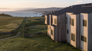 A vibrant dog park bathed in sunlight features abundant greenery with mature trees, offering ample shade for leisurely outdoor activities. In the distance is a well-equipped playground that presents an additional activity area. An inviting picnic table spreads out nearby, ideal for enjoying meals al fresco or taking relaxing breaks from play. A neatly-placed trash bin aids in maintaining the park's cleanliness for everyone's enjoyment. Dominating the scene is a generously wide dirt path gracefully curving through the expanse of grass, promising exploratory walks and lively fetch games.